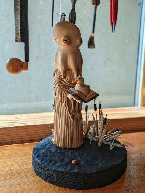 Woodcarved scene of a humanoid octopus wearing a dress on the surface of a pond. The creature has a kingfisher on its head. It's holding a book wich has a duck sitting on it. On the left of the base looking like water, bulrushes. On the right, a little frog has its head out of water. In the background woodcarving tools.