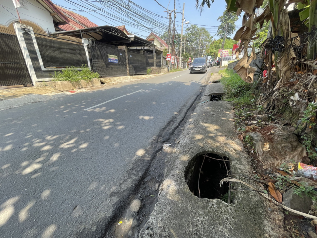 A suburban street with a large, uncovered holes on the sidewalk next to the curb. The holes are over a drainage system. The street is lined with residential houses, and electrical wires are visible overhead.