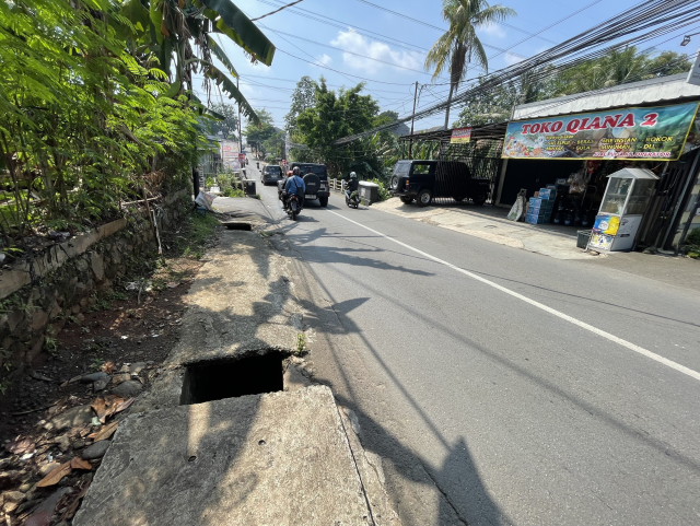 A suburban street scene showing multiple uncovered drainage holes along the sidewalk. A motorbike and cars are passing by, and a bodega called "Toko Qiana 2" is visible on the right side of the road. There is greenery on the left side of the street, including banana trees.