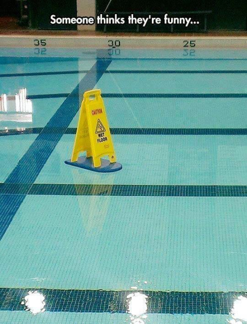 A yellow plastic A-frame warning sign floats on a kick-board in the middle of an indoor swimming pool. The sign reads: CAUTION 
WET FLOOR

Caption: Someone thinks they're funny... 