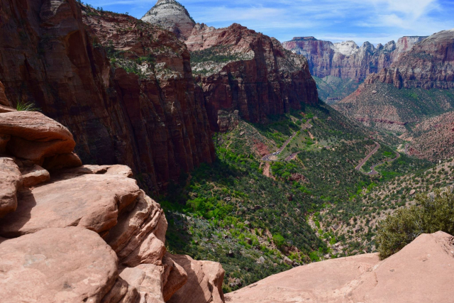 Zion National Park. Scenic view of a canyon with rocky cliffs, a winding road, and a lush valley, under a blue sky with scattered clouds.