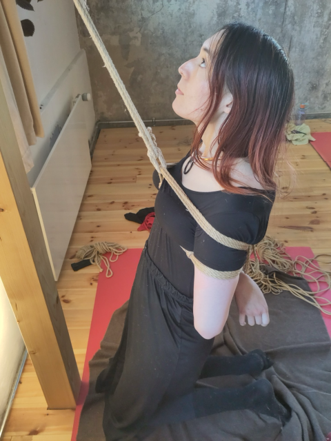 Elise kneeling on the ground facing the hashira with a rope from an anchor above on the hashira out of frame attached to a harness tied around her chest, squeezing her breasts together from the top and bottom, and her arms tied behind her back. She is looking up at the hashira 