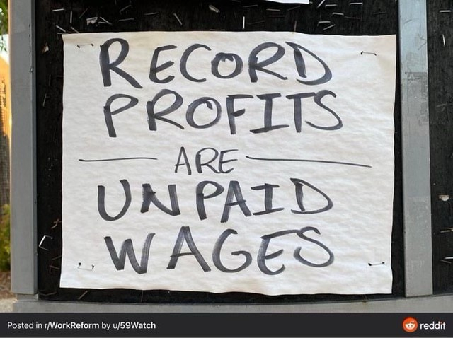 A handwritten sign reads "RECORD PROFITS ARE UNPAID WAGES." The sign is posted on a wall.