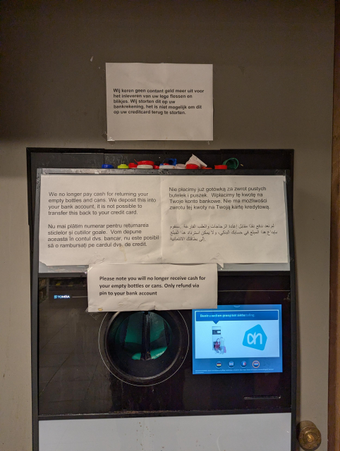 A TOMRA reverse vending machine  covered with several signs in many languages.
The bottom most sign reads: "Please note you will no longer receive cash for your empty bottles or cans. Only refund via pin to your bank account"