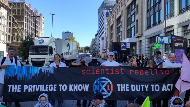 A group of protesters blocks a street in Brussels. A couple of them, wearing white lab coats, are holding a banner saying 'Scientist Rebellion. The privilege to know, the duty to act'. The entrance to metro station 'Rogier' is visible in the background.