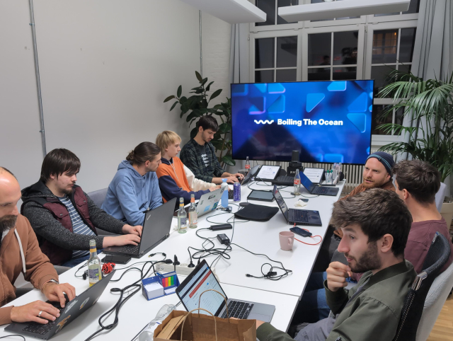 About 10 hackers sitting at a long conference table with their laptops. At the end of the table there's a large TV with a GNOME wallpwaper and the text "Boiling The Ocean". Behind the TV there are some plants.