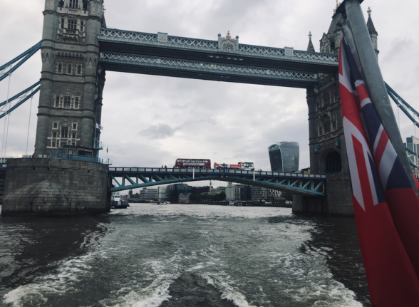 Photo shot from a boat that's just passed through Tower Bridge, leaving a churned-up wake. In the distance, a big red London bus crosses Tower Bridge. 