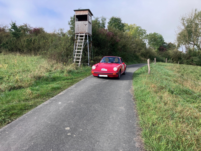 Roter Porsche fährt auf geteertem Feldweg neben einem Hochsitz.