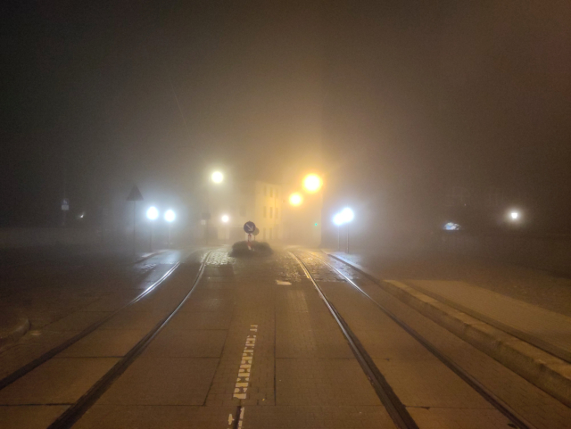 A very foggy street by night. Two tram tracks are visibile as well as blurry white and yellow street lights.