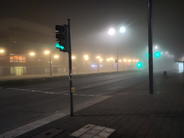 Another foggy street. In the background blurred green traffic lights are visible through the fog.