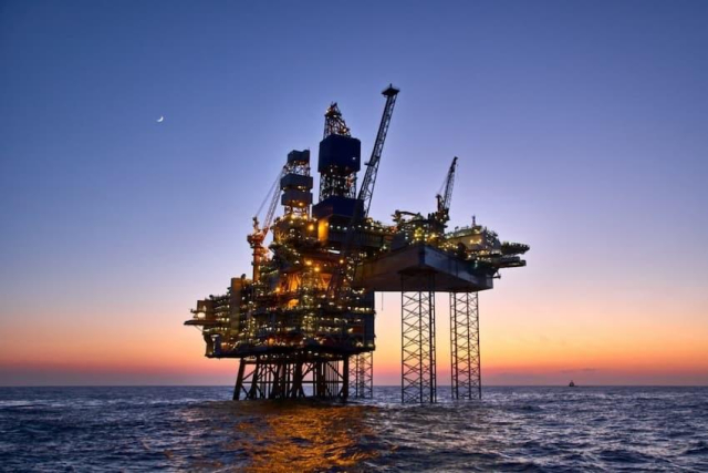 An offshore oil platform illuminated against a twilight sky over the ocean, with cranes and support structures visible.