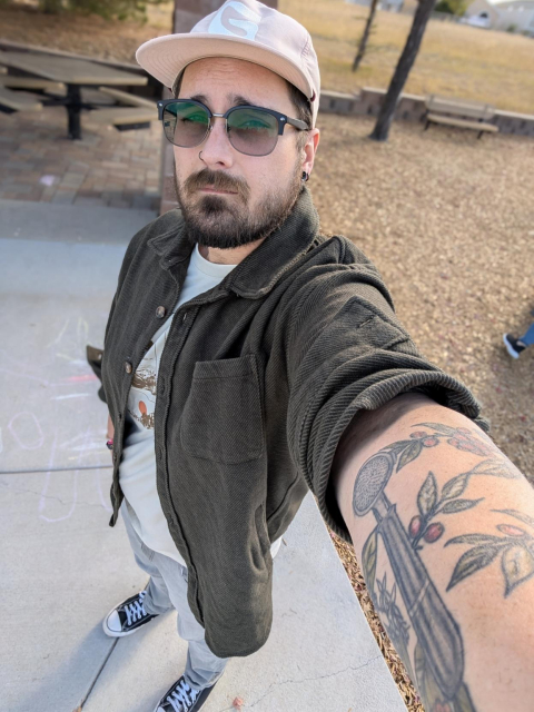 Selfie taken with the wide angle lens. I'm wearing a thick green flannel button up with the sleeves rolled up, a off white graphic T-shirt, grey wash jeans, and black low top converse shoes. You can see my tattooed arm that's holding the camera and it makes for a kinda cool perspective thing.