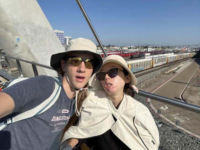 Us on the bridge wearing hats and sunglasses. I'm wearing a Dongletown shirt. The combined freight and trolley yard is behind us, with red trolleys, tank cars, and tall cars with corrugated metal walls and rounded roofs.