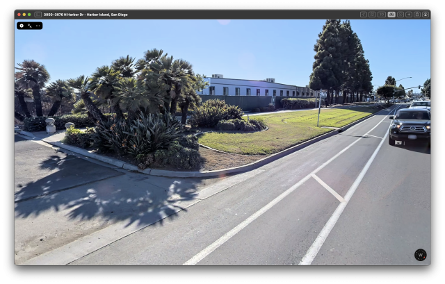 A screenshot of Apple Maps Look Around, showing the bus stop, which is indicated by a sign sticking out of a concrete storm drain. No sidewalk connects to where this sign is, because it is about 4 meters behind the sign, separated from the road by grass. At the left side of the photo is the driveway to an industrial property, which is where we tried to wait for the bus. A non-separated bike lane is at the side of the road, which has a speed limit of 45 mph.