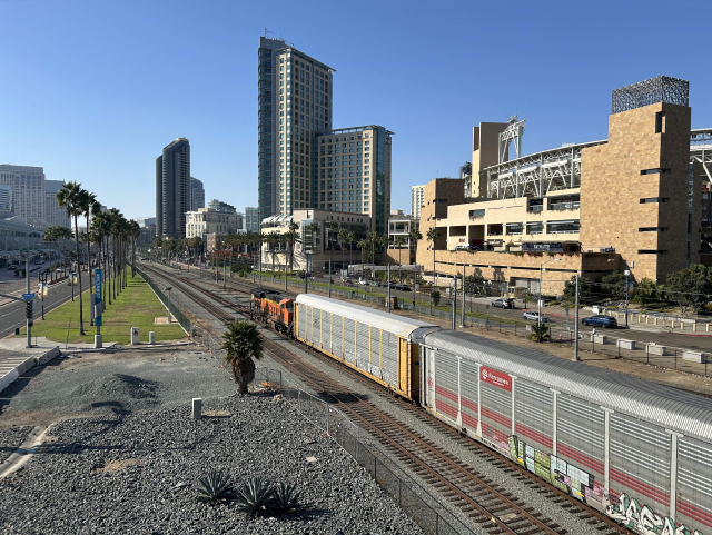 A view facing northwest from the pedestrian bridge, looking along the two trolley tracks and two freight tracks before they merge and enter downtown. A BNSF freight locomotive sits at the front of a line of corrugated freight cars, one of which says "Ferromex grupo Mexico" on its side. Adjacent to the bridge is a spot where an at-grade crossing clearly used to be, as the roadway is blocked off on the southwest side with concrete jersey barriers. A mix of medium and high rise buildings lines the inland side of the railway. The San Diego Convention Center can just barely be seen at the left side.