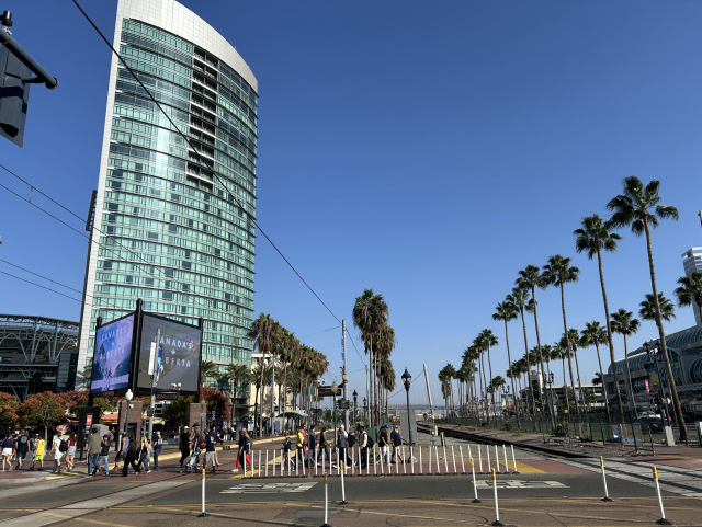 A view looking southeast from a point at a grade crossing between the two trolley tracks and the single main line freight track as they pass through the Gaslamp Quarter. Palm trees line both sides of the railway and the space in between the trolley and freight tracks. A collection of white plastic sticks delineate where pedestrians and vehicles are supposed to cross the tracks. A large crowd of people are walking across the trolley tracks toward the left side. A tall building sits to the inland side, to the left of the trolley tracks, with large LED displays toward its bottom.