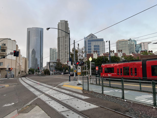 A view across the complicated intersection of Kettner Blvd and G st in downtown San Diego, where the two trolley tracks and one freight track curve roughly diagonally through the middle of the road intersection. A trolley approaches the intersection. Tall buildings are in the background and the sky is overcast.