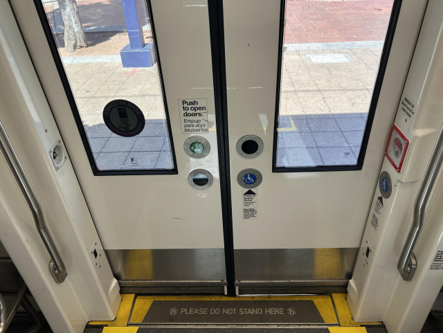 The inside of the lower 60% of a doorway on the trolley. At the center edges of the double doors are a series of buttons. The top left button has a label above it that says, "Push to open doors. Empuje para abrir las puertas." The bottom right button has a wheelchair symbol on it and a label below it that says, "Press to deploy ramp. Presione para extender la rampa. Doors must close before ramp deploys. Puertas deben cerrar antes para extender rampa." The top right and bottom left buttons are blank circles. The threshold, which slightly slopes down, has text that says, "Please do not stand here."