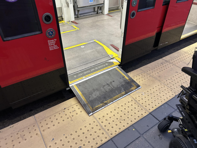 A ramp extending out from the threshold of a trolley door onto the yellow bumpy platform. The door is open. The button with the wheelchair symbol on the door has a ring of red lights lit up around it. Part of the bottom and front of Aubrie's power wheelchair is visible at the bottom right of the photo, approaching the ramp.