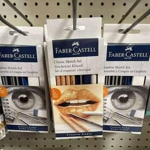 The image shows three boxes of Faber-Castell Classic Sketch Sets. The boxes are on a shelf in a store. The boxes are blue and white with a picture of a sketch on the front: one eye on the left , one mouth on the middle, and one eye on the right.The text on the boxes is in English, German, and French. The text on the boxes says that the set contains 12 graphite pencils, 1 eraser, 1 pencil sharpener, and 1 blending stump.
