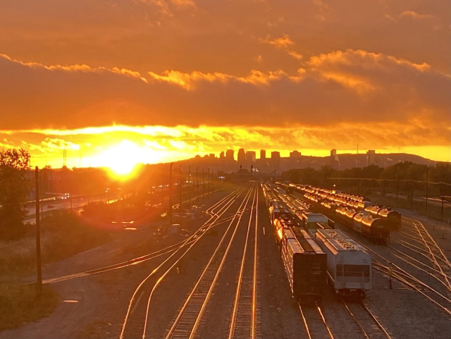 A stunning sunset with a bright yellow orb sitting on the horizon, making the sky all around it bright yellow, then blending into orange. In the foreground are train tracks and train cars that are reflecting the golden sun, and on the horizon is the Montreal skyline. 