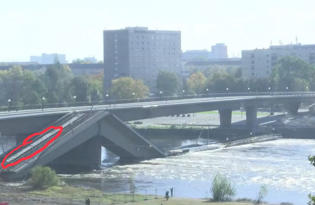 Screenshot des Livestream des MDR von der CarolaBrücke in Dresden. Auf dem man die Elbe sieht und Teile der eingestürzten Brücke sowie im Vordergrund, einen Bauzaun unter dahinter Menschen, die sich für die CarolaBrücke interessieren. Auf einem der Teile der Carola Brücke ist undeutlich ein Graffiti zu erkennen. Dies habe ich rot eingekreist.