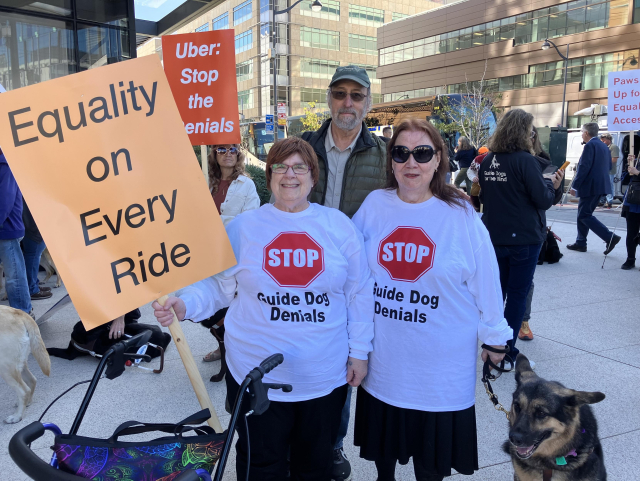 Photo of Kim Charlson (right) and her guide dog Idabelle standing with Vicki Vogt (left) from the Perkins School for the Blind and Lukas Franck (center) from Vision Loss Alliance of New Jersey. Kim and Vicki wear white t-shirts that say “STOP Guide Dog Denials.” Vicki holds a sign that reads “Equality on Every Ride.” They stand in front of an office building with other people at the Rideshare Rally. 