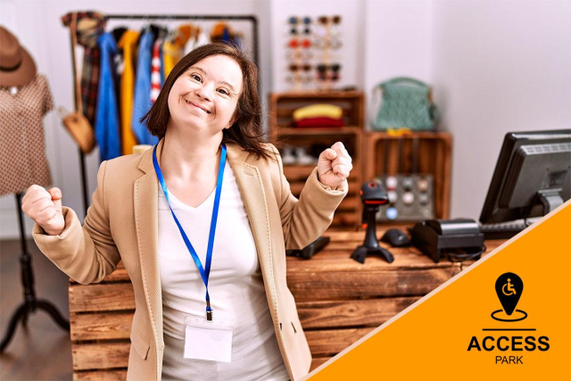 A young woman with Down syndrome is smiling brightly and raising her fists in a celebratory gesture, as if excited or triumphant. She is wearing a beige blazer over a white shirt with a blue lanyard around her neck. Behind her, various clothing items are displayed on racks, and shelves hold neatly folded clothes, suggesting she is in a retail or boutique setting. In the foreground, a computer monitor is visible on the right, indicating she may be working at the store or assisting customers. The overall mood is joyful and positive. Bottom Right: A simple but iconic black logo features a map pin icon with a wheelchair symbol inside it, pointing downward to a place of accessibility. To the right of the pin, there is vertical line separating the icon from the bold uppercase word 'ACCESS,' with the word 'PARK' written in smaller capital letters below. The design suggests a focus on 'Access Park,' where access if provided for all.