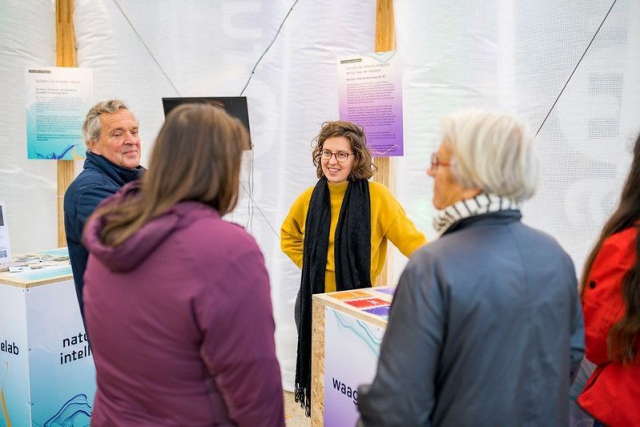 Een groepje mensen staat in een witte tent. Eromheen zijn borden geplaatst in turquoise en groen. Een vrouw met een gele trui, bruin haar en bruine bril kijkt lachend naar een andere vrouw die staat te spreken met haar rug naar de camera gekeerd.