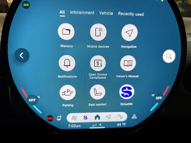 Part of a circular infotainment screen of a vehicle, showing a 3x3 grid of categories as round white icons, all on a blue background. The text includes:

Header tab (All  Infotainment  Vehicle Recently used) with "All" selected.

The labels underneath the icons:
Memory  Mobile devices  Navigation
Notifications  Open Source Compliance  Owner's Manual
Parking  Seat comfort. SiriusXM
