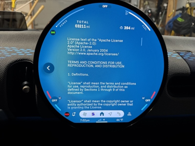 A circular vehicle infotainment screen. It shows the full text of the Apache License 2.0 open source software license in white lettering on a blue background.

Behind the round screen part of the vehicle dashboard is visible.