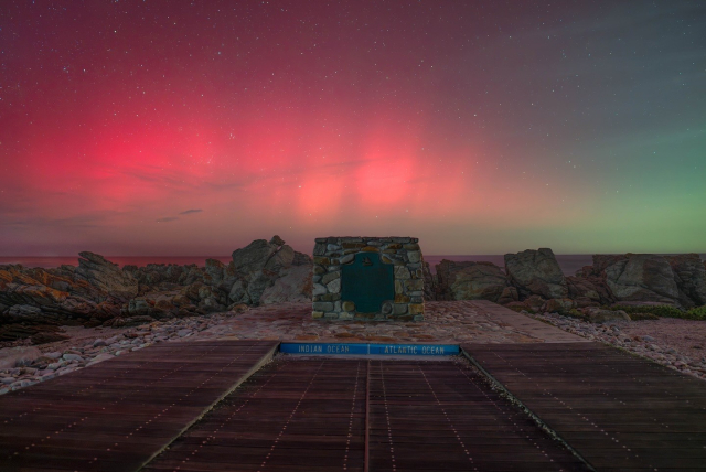 This image shows a striking natural phenomenon at the meeting point of the Indian and Atlantic Oceans, marked by a stone monument in the foreground. The sky above is illuminated with hues of red, pink, and green, suggesting an aurora display, which creates an otherworldly atmosphere. The rugged rocks and ocean in the background add to the dramatic scene, emphasising the wild beauty of this unique location.