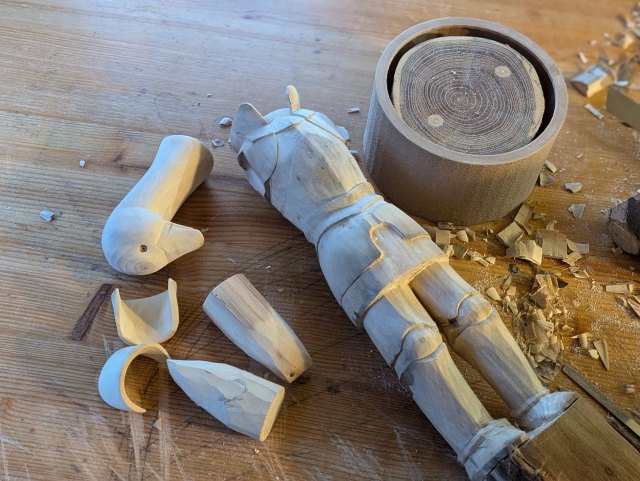 Pieces of the draft of a woodcarved humanoid goose in plate armour on a workbench 