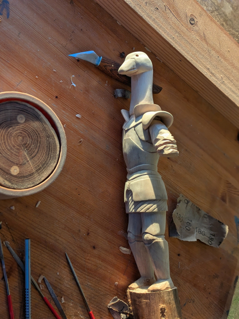 Draft of a woodcarved humanoid goose wearing plate armour. On a workbench