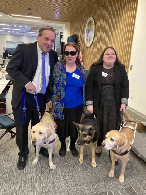 Picture of Carl Richardson and his guide dog Dayton (left), Kim Charlson and her guide dog Idabelle (center) and Claire Stanley and her guide dog Tulane (right). The group is standing in a meeting room at the FCC. 