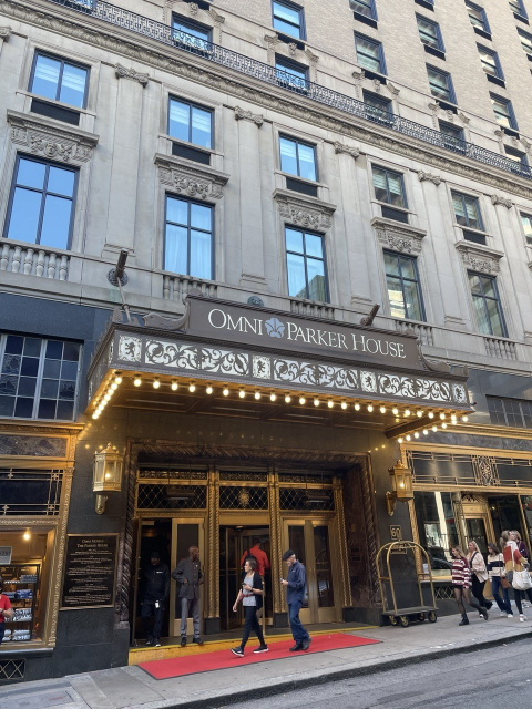 A grand 19th-century style building entrance, with a red carpet in front and a few pedestrians and uniformed door staff visible. The sign over the lightbulb-ringed awning reads OMNI PARKER HOUSE HOTEL.