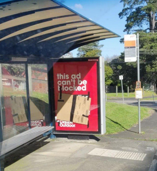 a bus station during a sunny day, which has an advertising panel. on it there was an advertisement for outdoor, where it's written: "this ad can't be blocked". The text that follows can't be read because someone has covered it with a cardboard box, or an "adblocker" in the physical world.