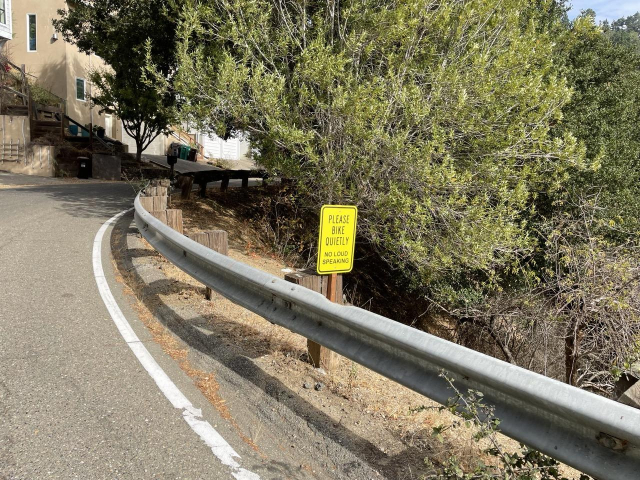 on the side of a hilly road with houses on the left side, a small yellow sign that says “please bike quietly - no loud speaking”