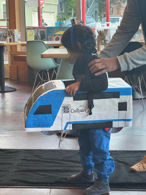 A small child, wearing a cardboard box decorated as a new Caltrain engine. The child also has a small headpiece, connecting them to invisible electrical wires.