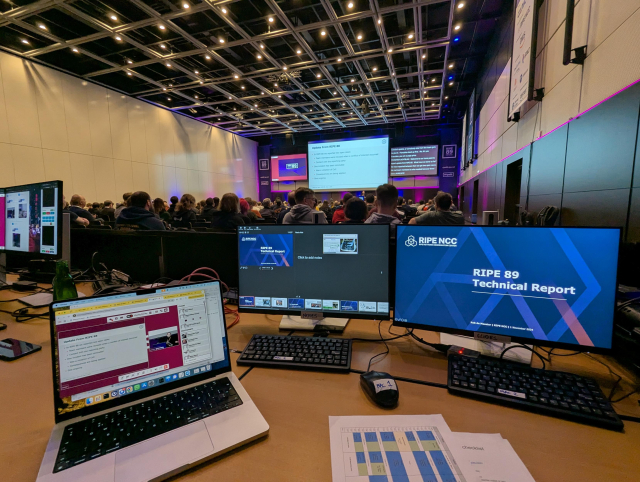 A photo of a room with RIPE 89 on screens in the front and some other presentation running in the background