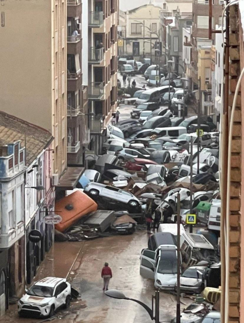 Photograph taken in Alfafar, Valencia, Spain, showing a city street littered with overturned and piled-up cars after massive floods caused by heavy rainfall. The road is covered in mud and debris, and a few people walk through the wreckage amidst buildings that appear largely intact. The scene highlights the extensive destruction and disruption caused by the flooding.