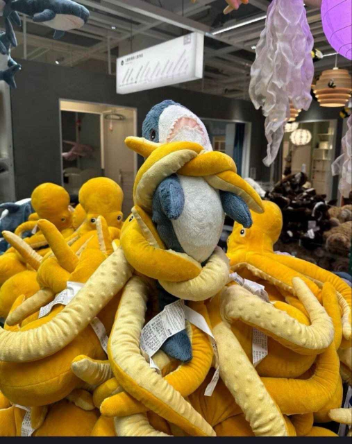 A store display. A blahaj, a blue and white shark plushie with a pink mouth, sits on top of a pile of beige and white plushie octopodes. Many of the tentacles are reaching up and wrapping around the blahaj. It's posed as though it's trying to escape their clutches.