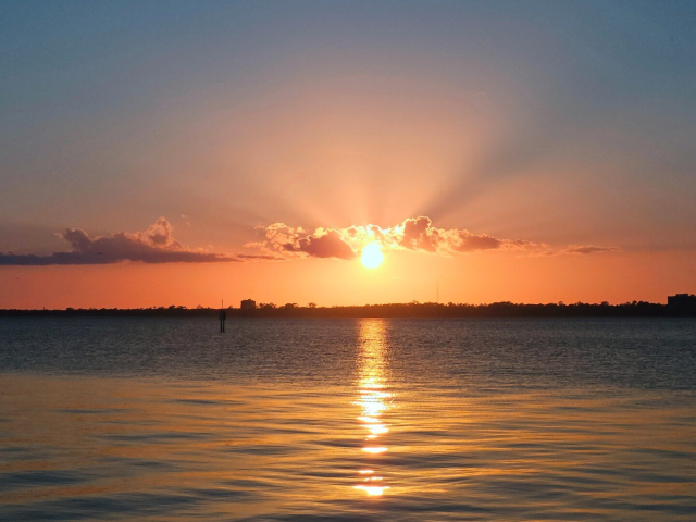 An orange glow illuminates the horizon over a wide, calm blue river. At the center, a bright, blinding sun gets caught between some small cloud formations which causes sun beams extended out in all directions. The warm glow reflects onto the water's surface where gentle ripples of movement.