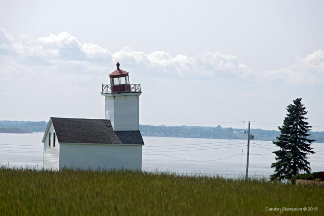 A white building with a white steeple on which the red-capped light housing was placed.  The red cap is tilted, having seen better days. 

In the foreground is a meadow, beyond is a still bay.  It's a bright, cloudy day.