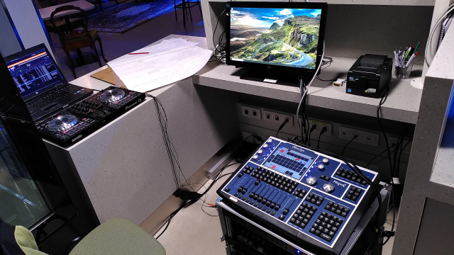 a counter top, on top of which are some papers, a point of sale system, my DJ controller and my laptop. next to the counter is a small moveable 19" rack on top of which is a DMX light control panel