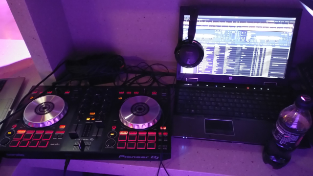 a DJ controller next to a laptop that is running DJ mixing software, there are headphones on the laptop. there is a mess of wires behind the DJ controller