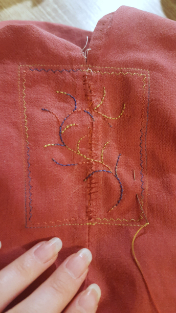 The stem or vane of whatever the embroidery will be, finished but with the fluffy bits or fronds missing. The shape is a lovely organic twist of curls, the thread colours stand out and it looks fairly balanced in the little framed rectangle of the topstitching holding the patch in place. The tear is visible when you look directly at it, but the eye is drawn to the embroidery, which will when finished cover up most of it.