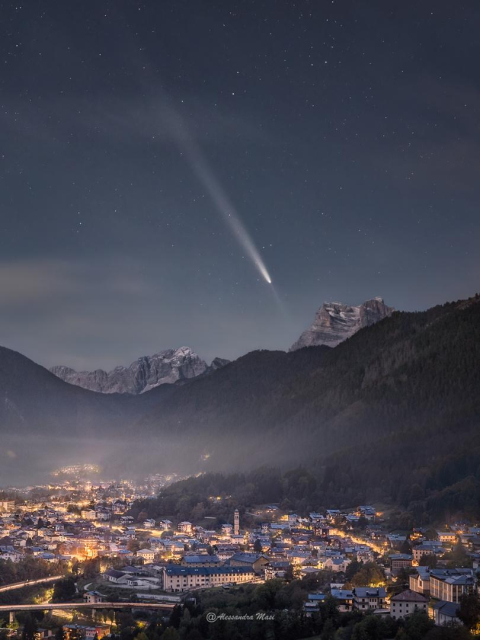 A town is shown in the foreground surrounded by tall mountains with even taller mountains in the distance. Above them all is a bright white streak that is a comet with both a tail and an anti-tail. High above are stars in the night sky.