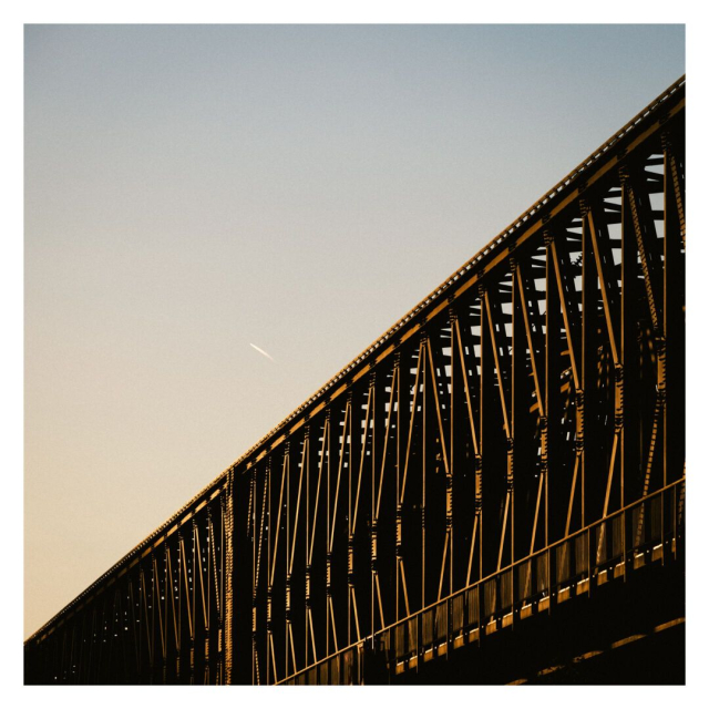 This is a square color image with a white frame, showing a bridge illuminated by the golden light of the setting sun. The bridge structure, which is composed of iron beams arranged in a repeating pattern, extends diagonally from the bottom left to the top right corner. The warm sunlight highlights the intricate details and casts shadows that emphasize the geometric shapes of the beams. The sky is a gentle gradient from light yellow to soft blue, and in the background, a small, subtle contrail is visible, adding a touch of movement to the otherwise still scene. The atmosphere of the photo evokes a calm, transitional moment as the day comes to an end.