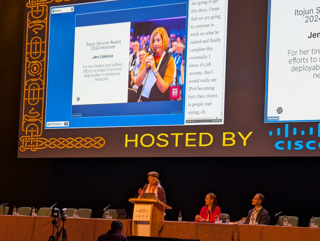 Jen Linkova at a podium in front of an IETF 121 plenary audience, accepting the award.